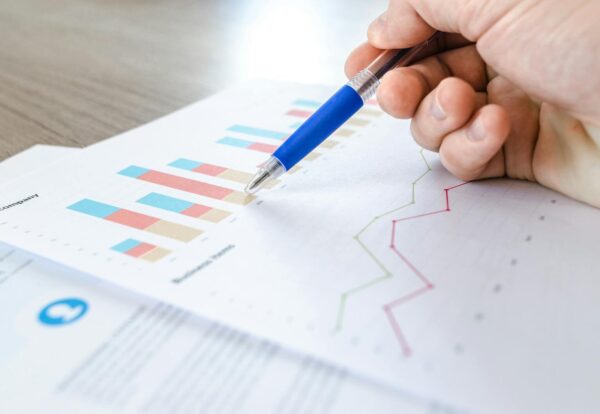 Photo by Lukas Blazek A close-up of a hand with a pen analyzing data on colorful bar and line charts on paper.