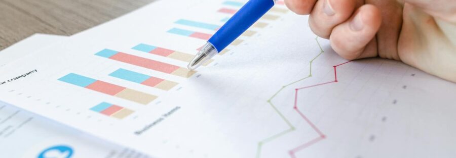 A close-up of a hand with a pen analyzing data on colorful bar and line charts on paper.